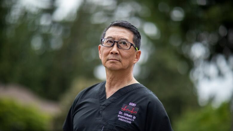 An Asian man wearing black glasses and black scrubs is pictured in front of a green hedge.