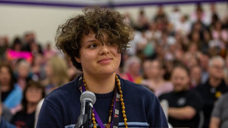 A young man with curly hair and wearing necklaces is standing behind a microphone. A crowd of people can be seen in the background.