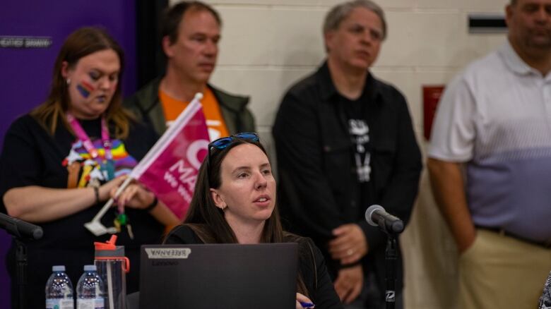 A woman is sitting behind a laptop. There are people standing behind her. 