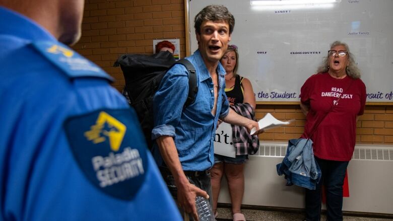 A man in a blue shirt is speaking to someone out of frame. A shoulder can be seen in the lower left corner, with a security guard badge on it. 