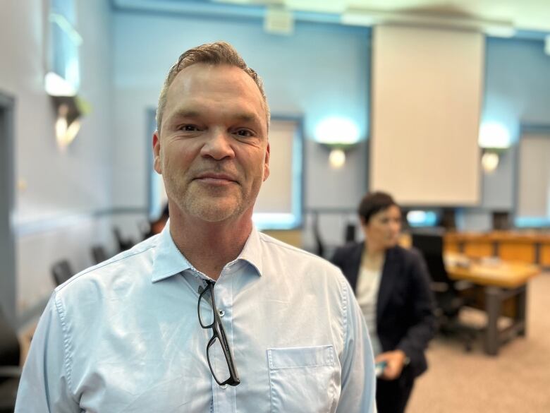 A man in a blue shirt with a pair of eyeglasses hanging from it standing in a mostly empty committee room