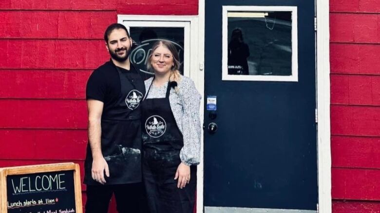 A man with a beard and a woman with a blonde ponytail stand in front of a red building. They are wearing black aprons that say 'White Sails Bakery and Deli'