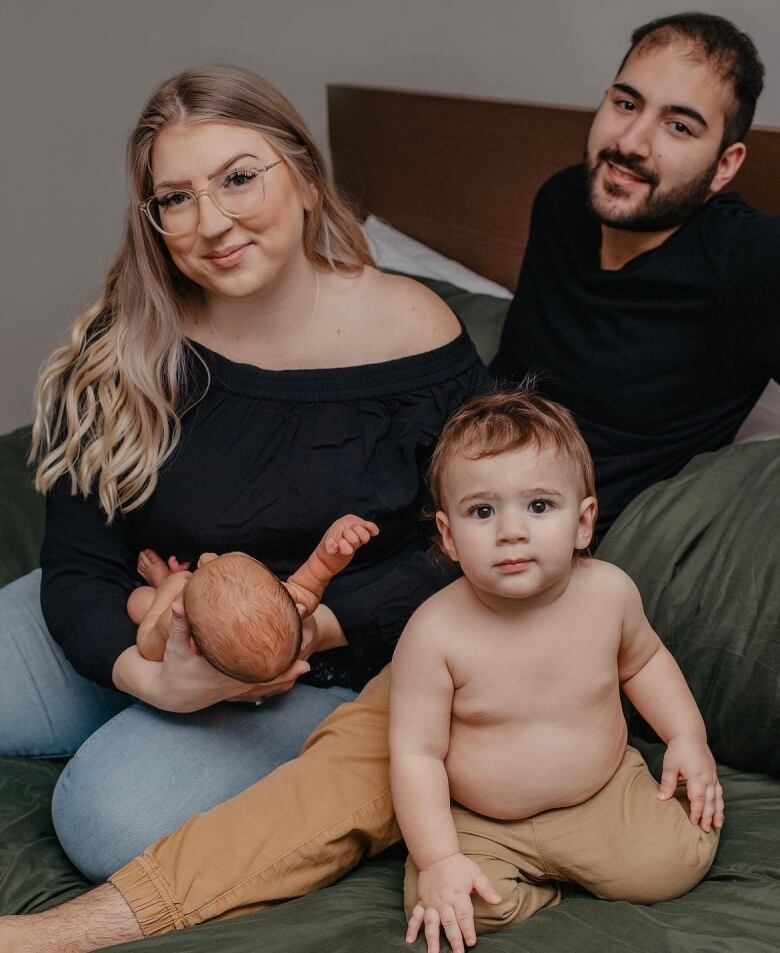 A woman with blonde hair and glasses and a man with a brown beard pose with their two kids. One is a toddler and one is a baby. 