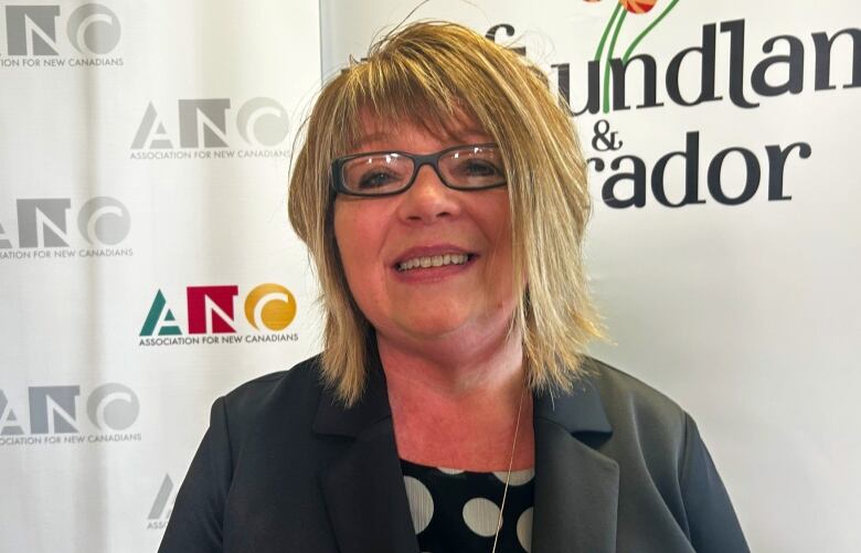 A woman with blonde hair and glasses smiling. Behind her are two backdrops. 