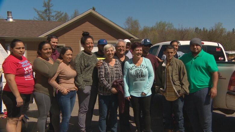 A group of 14 people pose for a picture in front of a pick up truck and a brown house.