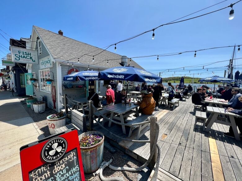 A patio with picnic tables is set up outside of a seafood restaurant. Some tables  are filled with people and have umbrellas overhead.