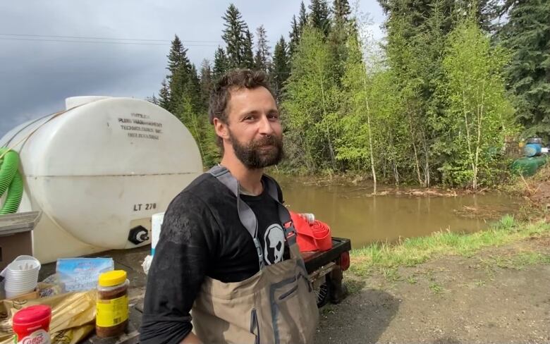 A man in bib overalls stands beside a plastic water tank with floodwaters visible behind him.