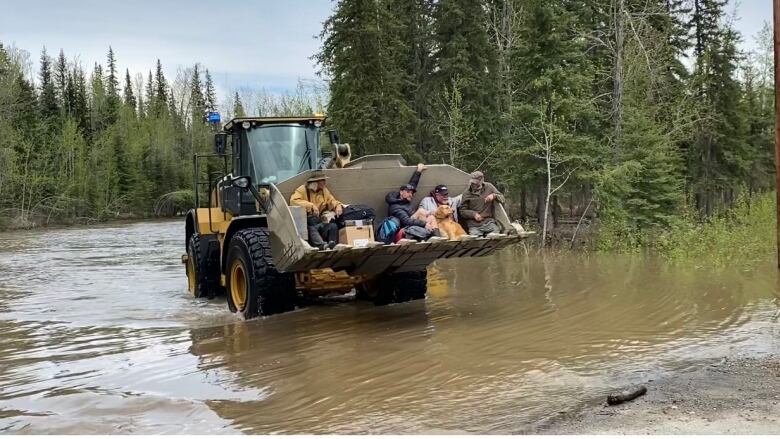 Four people sit in the bucket of an excavator driving on a flooded roadway.