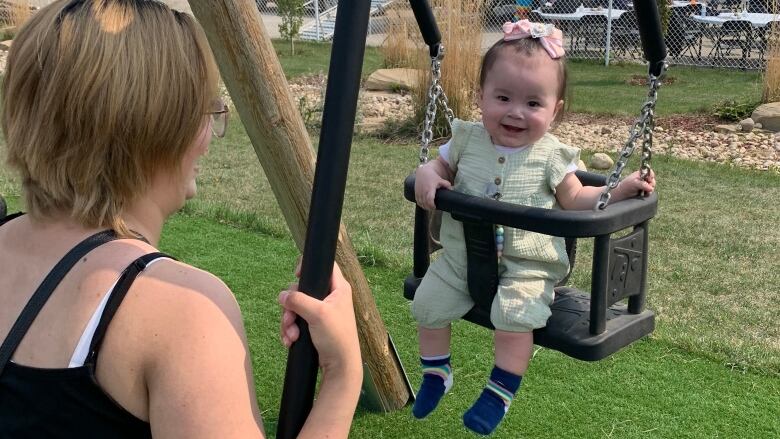 A mom sits on a swing facing her baby who sits on a smaller swing attached to hers at eye-level.