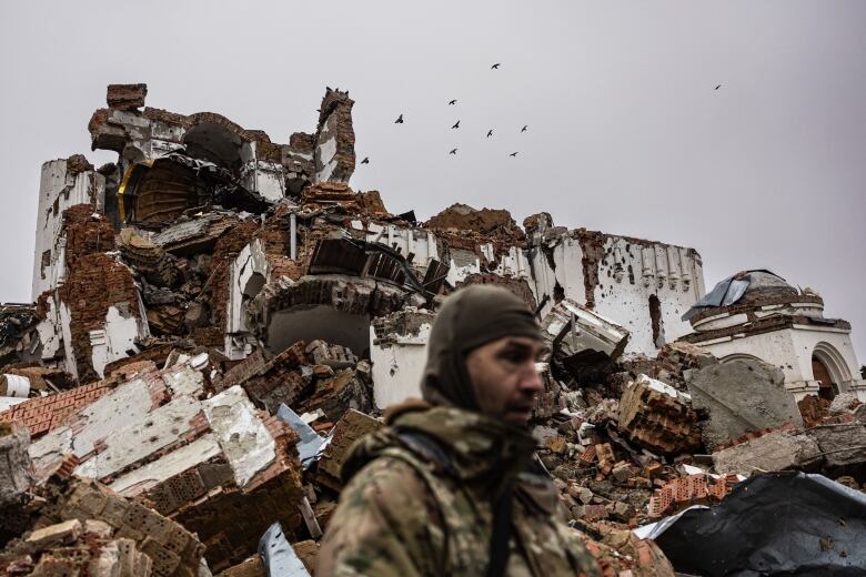 A man in a military uniform stands in front of the rubble of a destroyed building on an overcast day.