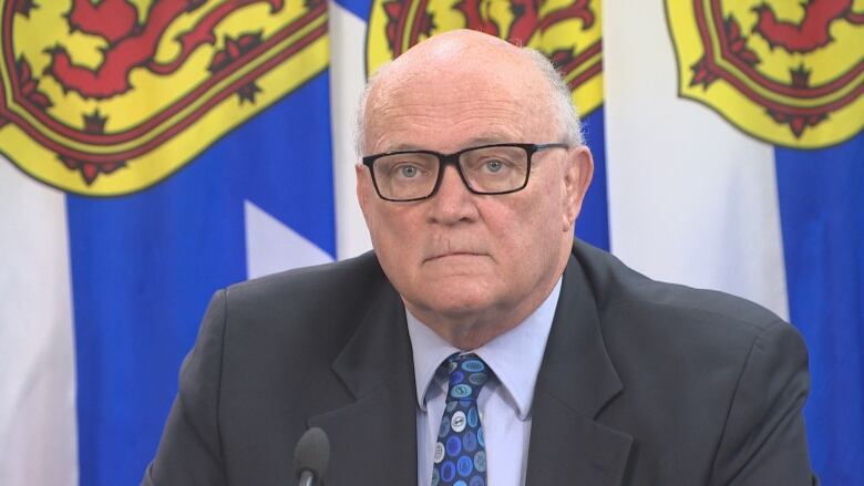 A man in a suit and tie sits at a table in front of a microphone with a Nova Scotia flag in the background.