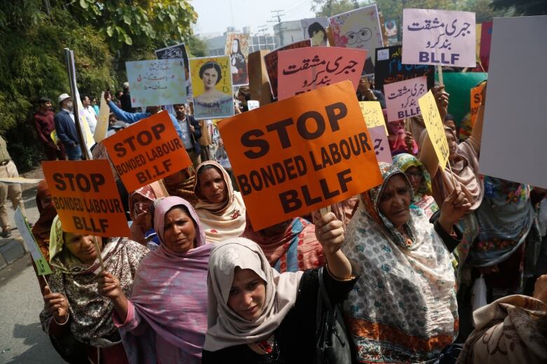 A group of Pakistani women wearing head scarves gather in a street holding orange placards that read Stop Bonded Labour BLLF. 