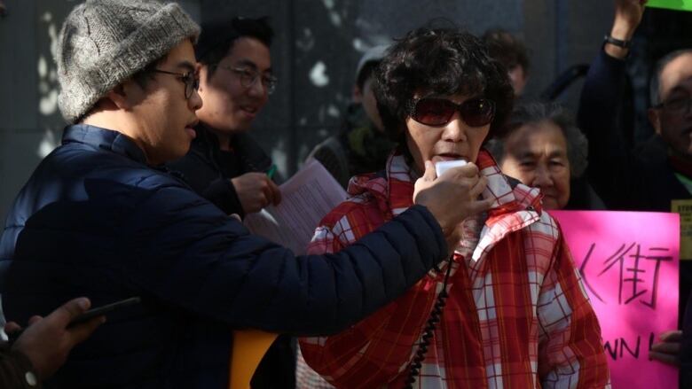 An Older Chinese woman speaks into a microphone at a protest outside Vancouver City hall.