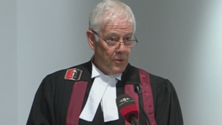 A man with white hair and glasses stands at a podium.