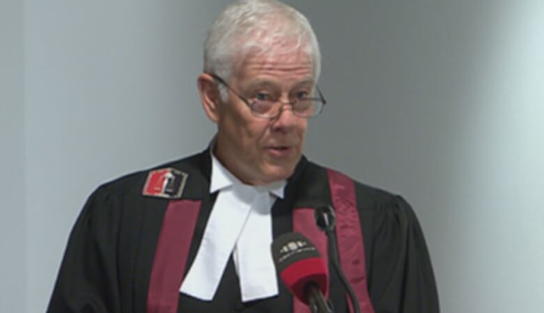 A man with white hair and glasses stands at a podium.