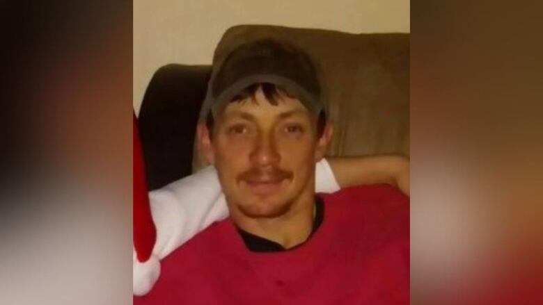 A man with facial hair wears a red long sleeve shirt and a black baseball hat.