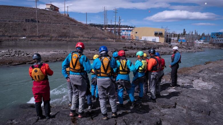 A group of people in lifejackets and helmets stand beside a fast-moving river in a city.