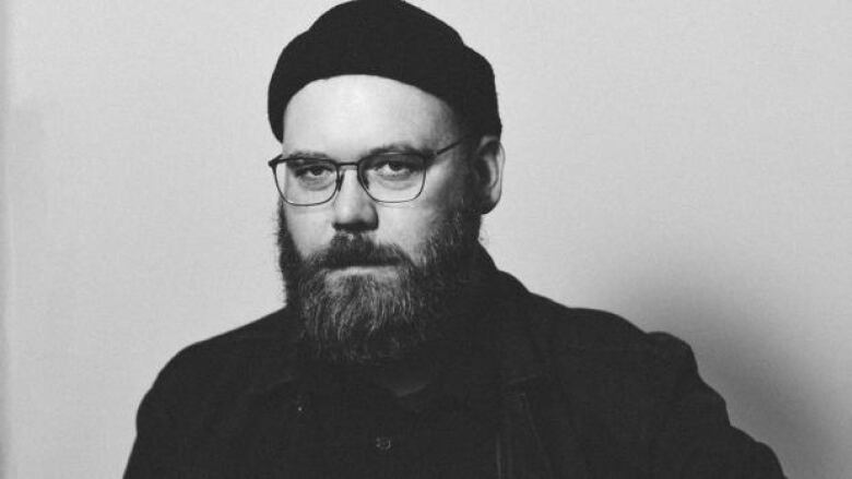 Black and white photo of a bearded man wearing glasses, a beanie, a black shirt and a dark jacket over it