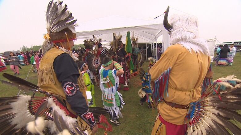 People of all ages are walking around on grass in traditional regalia at the James Smith Cree Nation 2023 powwow.