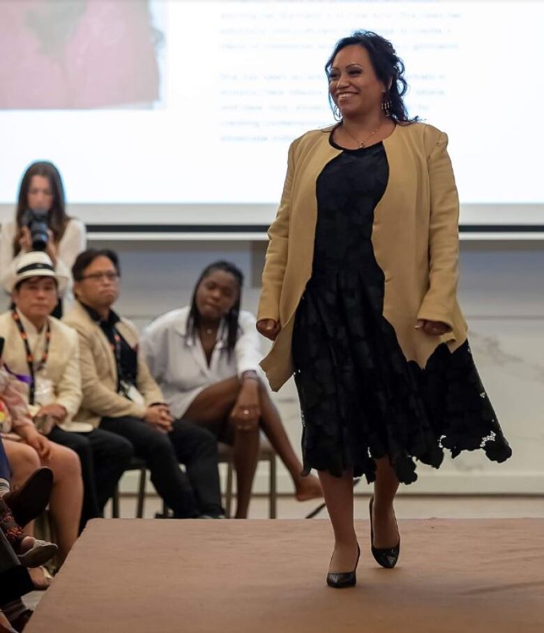 A smiling model in a tan jacket and black dress walks on a runway, with an audience watching.