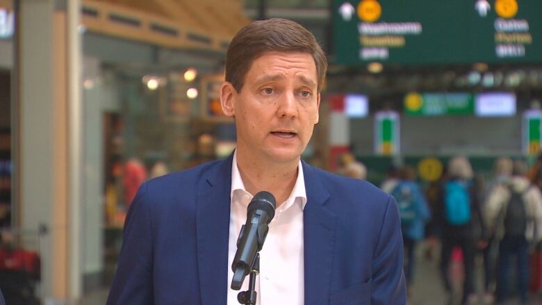 David Eby speaks into a microphone with the airport's interior in the background.