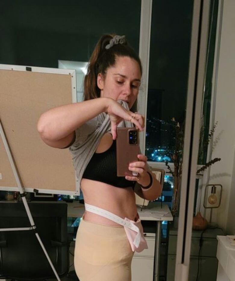 A woman exposes her stomach with a surgical pouch wrapped around it in her reflection in a mirror.