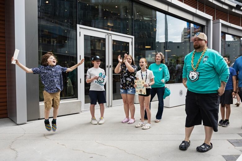 Four kids cheer with two holding signs saying 'go sea bears go', with one boy with medium length brown hair jumping in the air. Two adults dressed in blue green shirts with a bear logo look on.