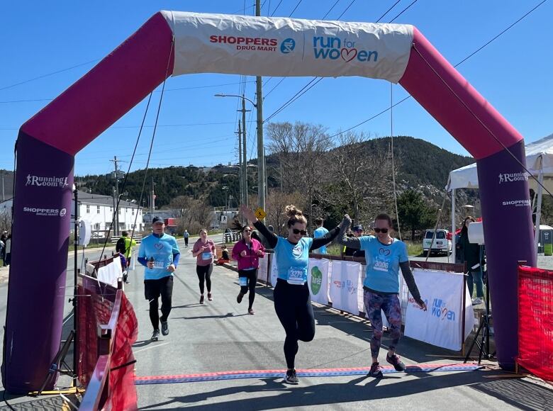 Two female runners are crossing the finish line and triumphantly hold hands in the air. Behind them more runners can be seen approaching the finish line.