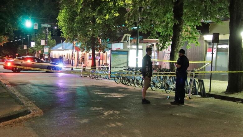 Police stand on a tree-lined street which is empty and blocked off by police tape.