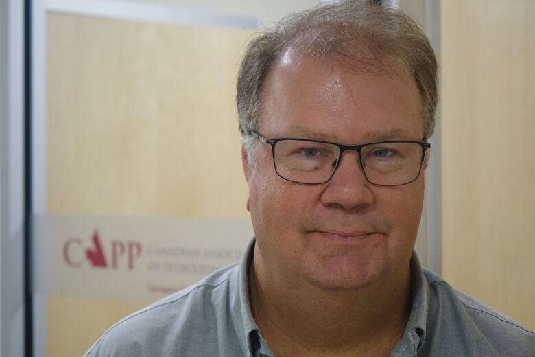 A man wearing a light green shirt and glasses stands before the office of the Canadian Association of Petroleum Producers.