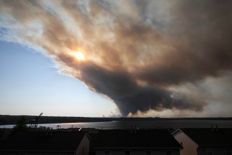 Across a body of water, plumes of heavy smoke drift upwards in the sky and spread outward from a wildfire in the distance.