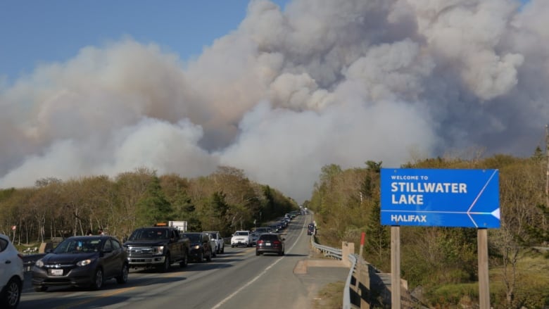 Cars on a road moving in the opposite direction of a wildfire whose heavy smoke can be seen in the distance in the sky.