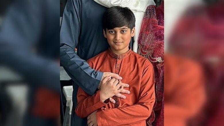 A young boy with short dark hair in a rust coloured tunic is seen holding his father's hand to his chest.