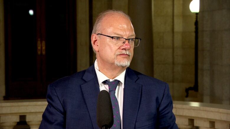 A white man in a navy suit and navy tie stands behind a microphone.