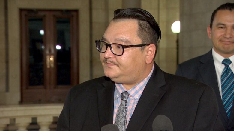 An Indigenous man with glasses wearing a black suit answers questions from reporters.