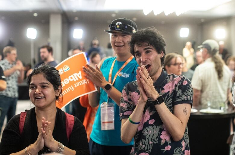 A woman and two men hold their hands in prayer as they watch Alberta election results come in.