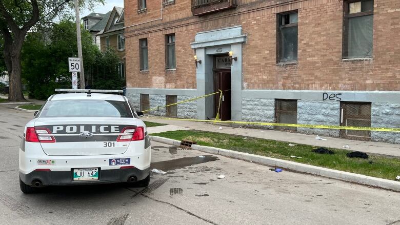 A black and white police car is parked on a street in front of an apartment building. Yellow police tape stretches across the front of the building.