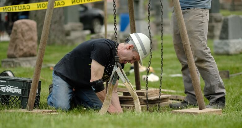 A man in a construction hard-hat kneels and reaches into a hole to steady a gravestone as it is winched up on chains connected to a supporting tripod.