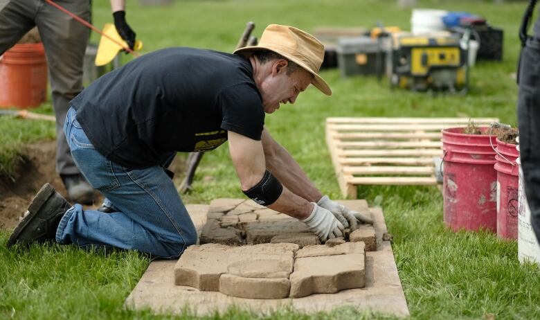 A man kneels on the grass and organizes the pieces of a broken headstone lying flat on the ground.