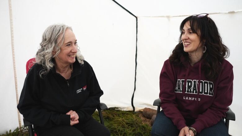 Two women sit on chairs on spruce boughs in a tent and smile at each other. 