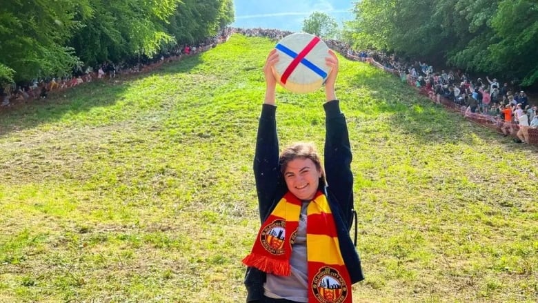 A woman holds a big wheel of cheese at the bottom of a hill