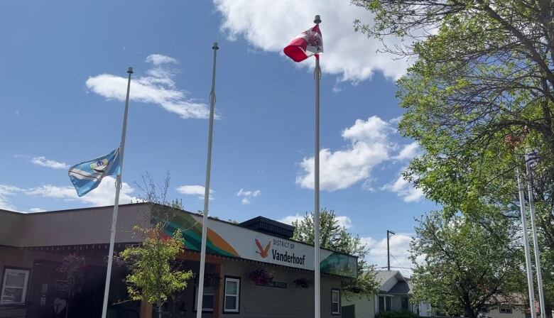 A flag at half mast outside a municipal office framed by a bright blue sky punctuated by fluffy clouds.