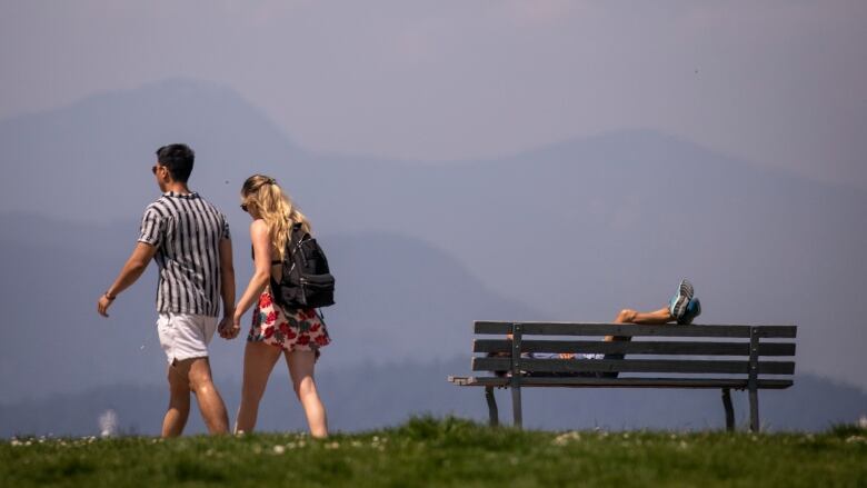 One person lies on a park bench while two people walk by amid hazy, smoky air seen in the mountainous landscape. 