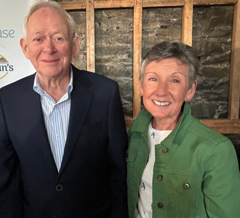 A man in a navy blazer and a woman in a green jacket pose in front of a stone wall with wooden beams. 