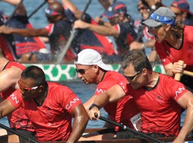 A female steersperson shouts from the back of a dragon boat at four male members of her crew, who are paddling vigorously with determination. 