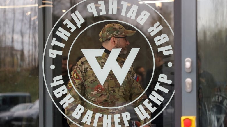 A man in a camouflage uniform stands behind a glass door bearing a white logo containing the letter W in the centre and encircled by words in Russian.