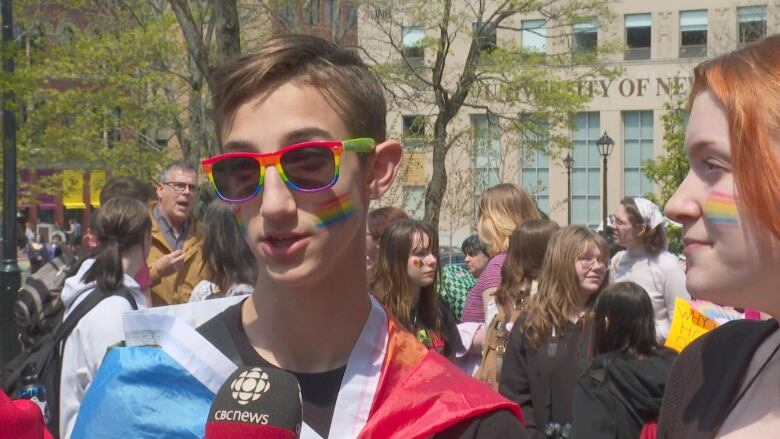 Boy wearing a rainbow flag as a cape, rainbow sunglasses and rainbow facepaint speaking into a microphone.