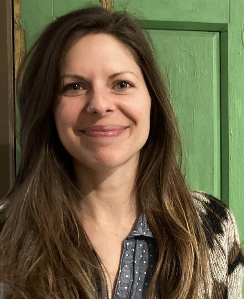 A woman with long golden-brown hair stands in front of a damaged wooden door that is painted lime green. She is wearing a dark blue-grey button up blouse with white polka-dots and a brown cardigan that has dark chevron strips around the top.