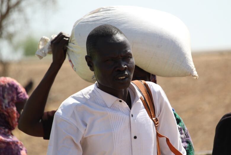 A tired looking man looks at the camera. A person carrying a sack on their head is behind him.