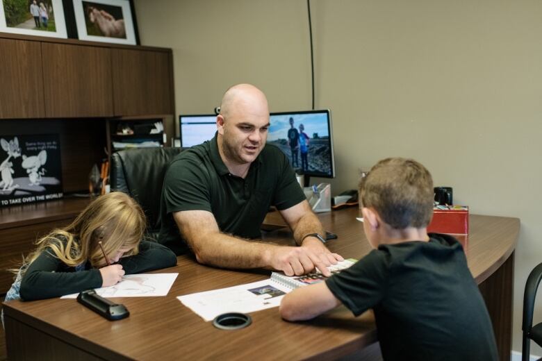 A man sits at a desk alongside two children.